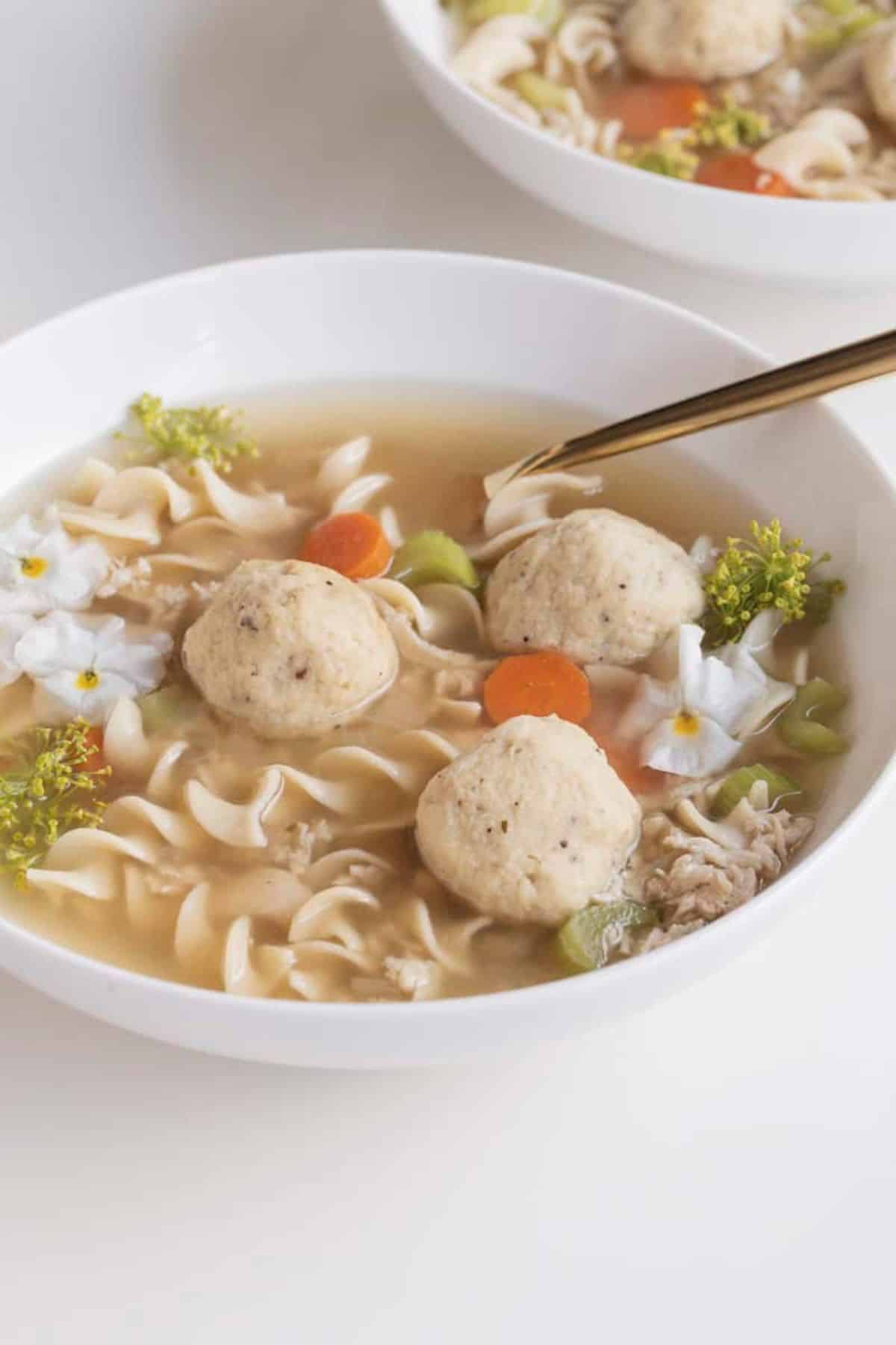 matzo ball soup in a bowl with noodles, chicken, veggies, and edible flowers