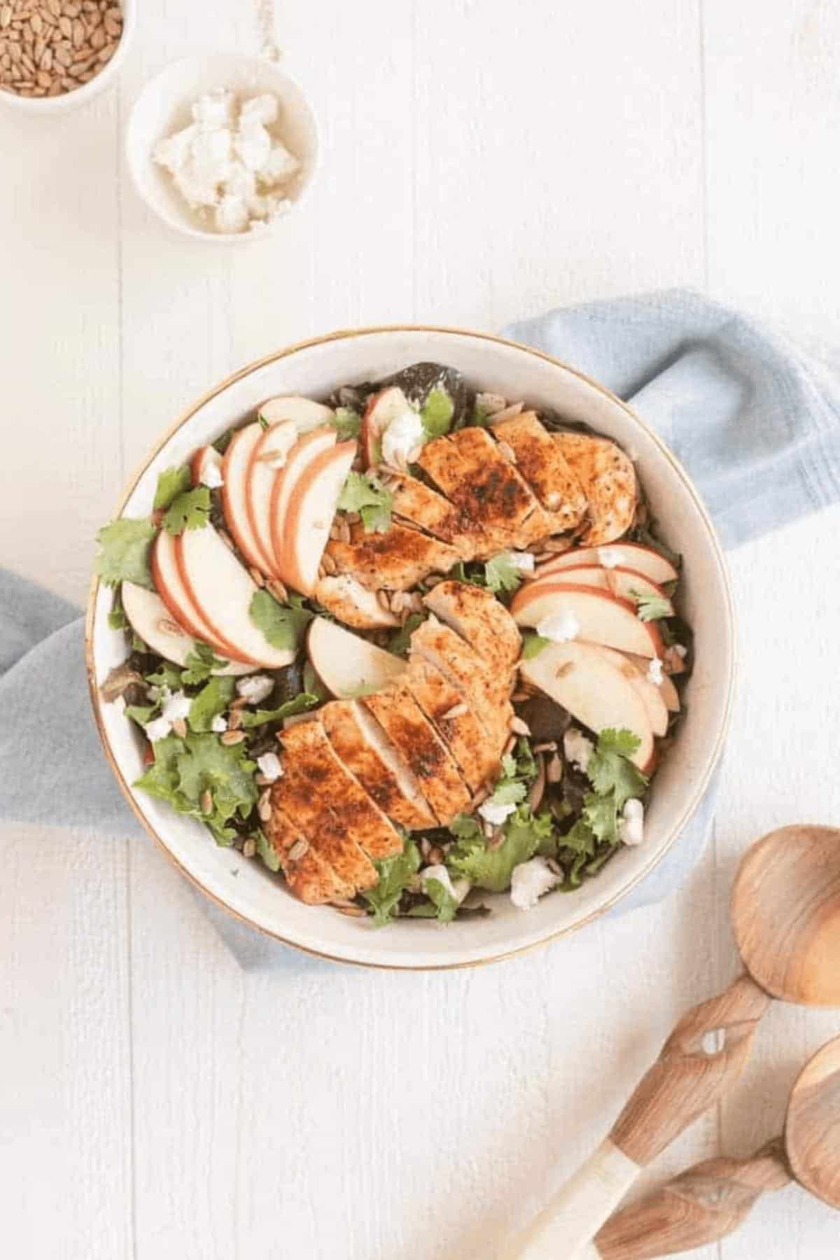 salad in a bowl with chicken and apples on top on a white surface